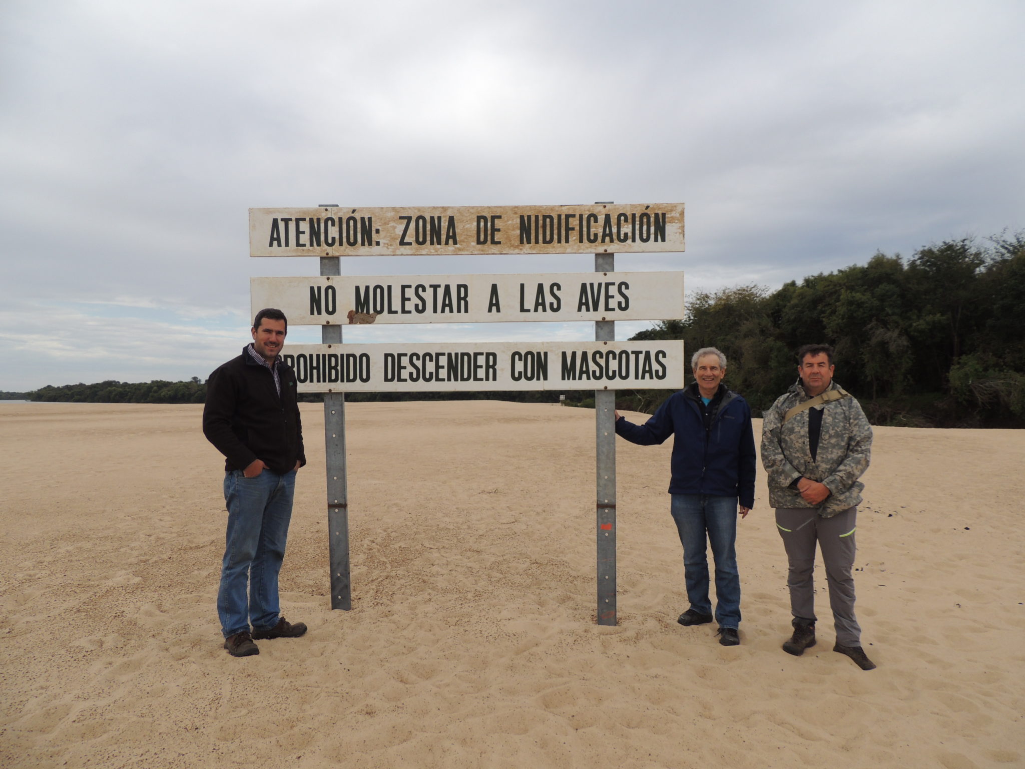 Three men stand near a tall sign on Queguay Island that warns tourists not to disturb nesting birds