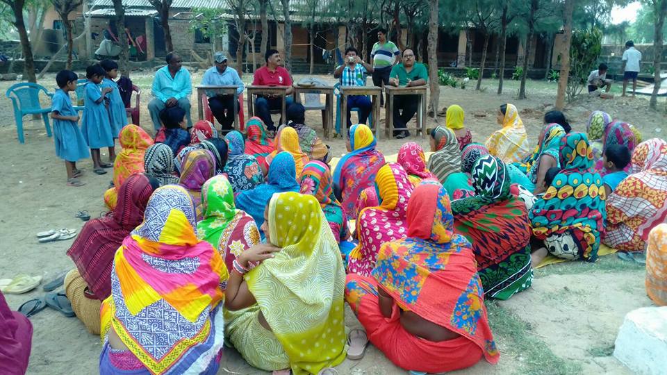 Women in saris sit on the ground for a meeting