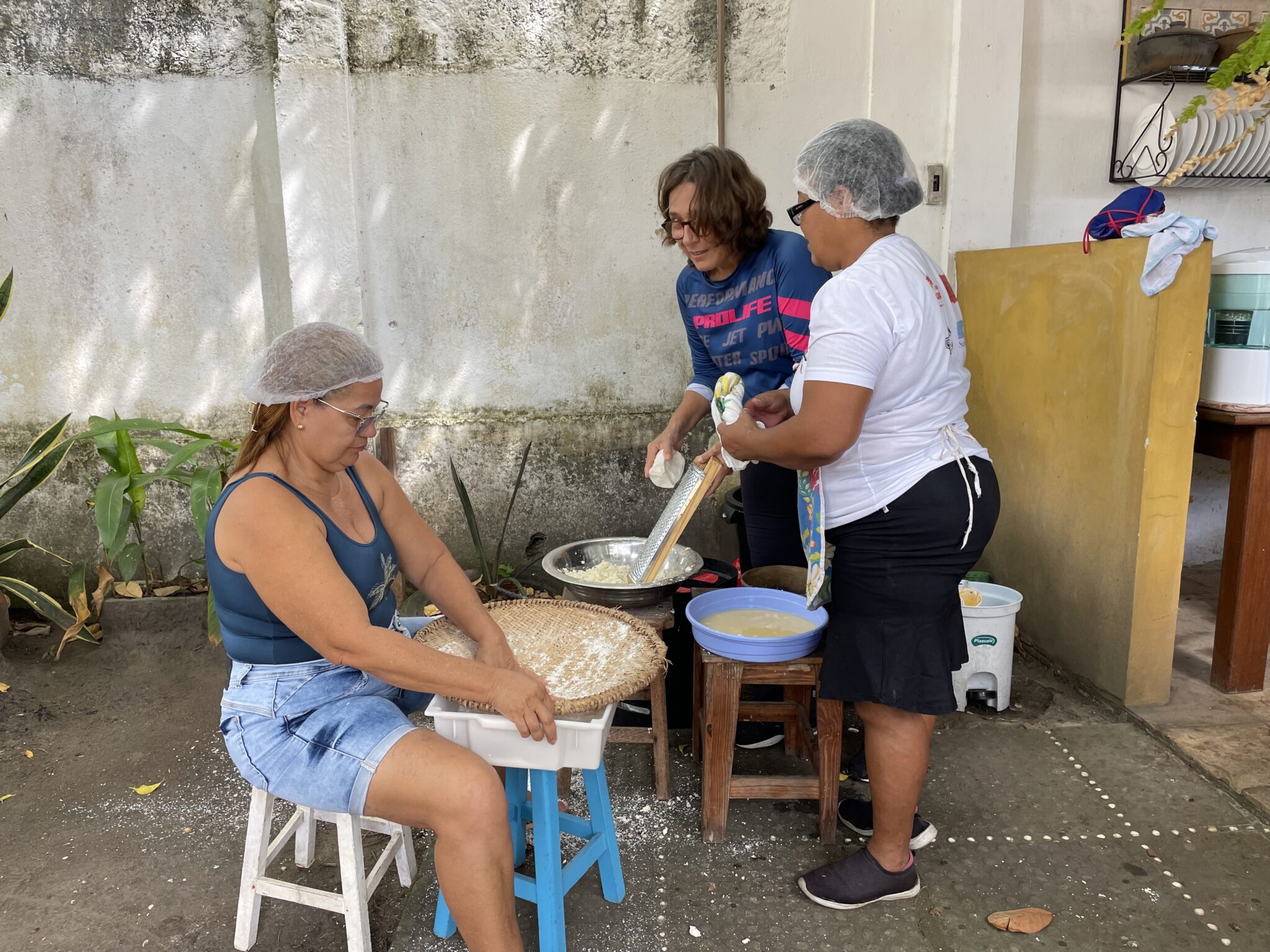 Three women stand or sit around cooking implements