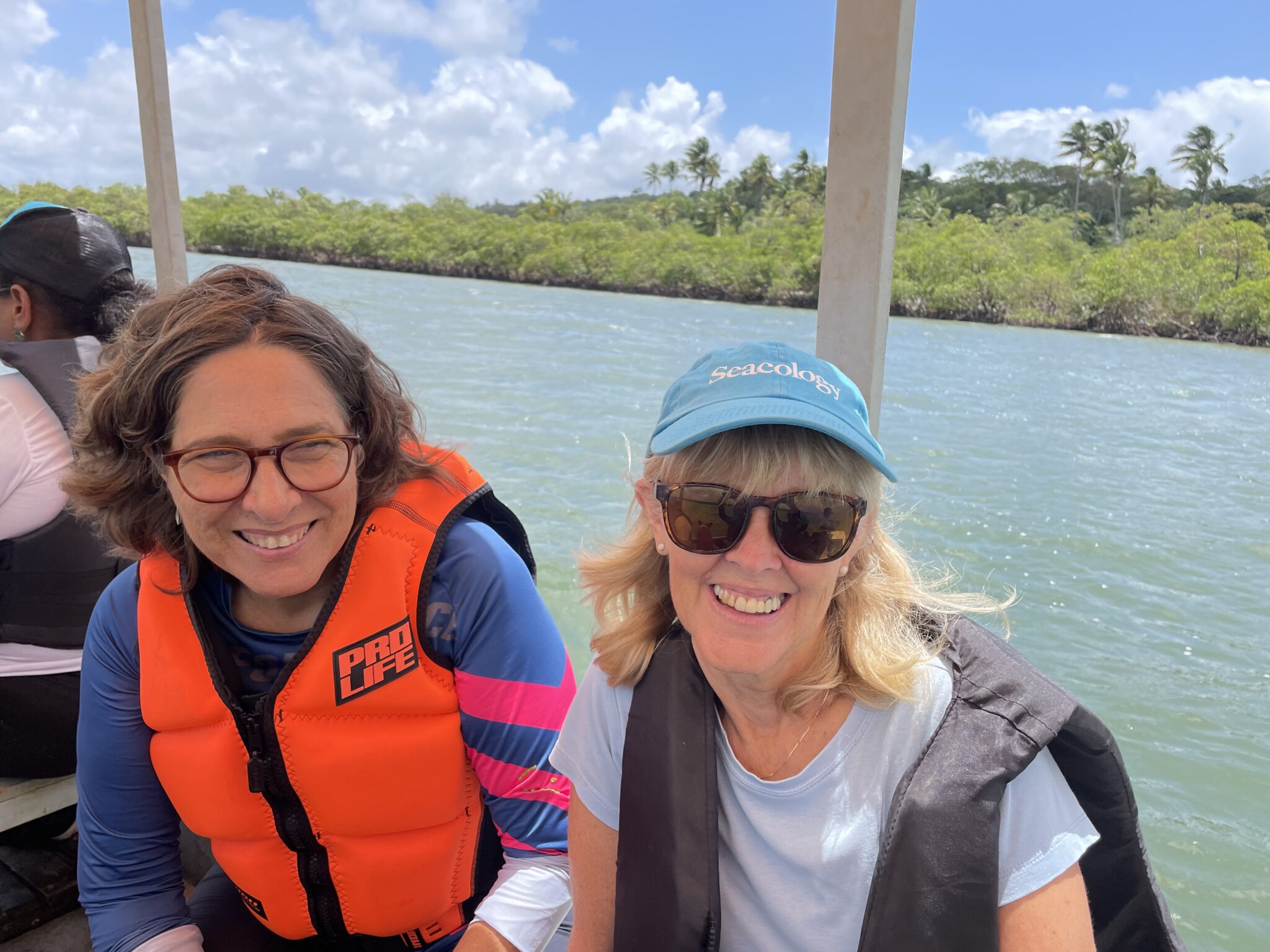 Two people sit on the side of a boat in a river and smile at the camera