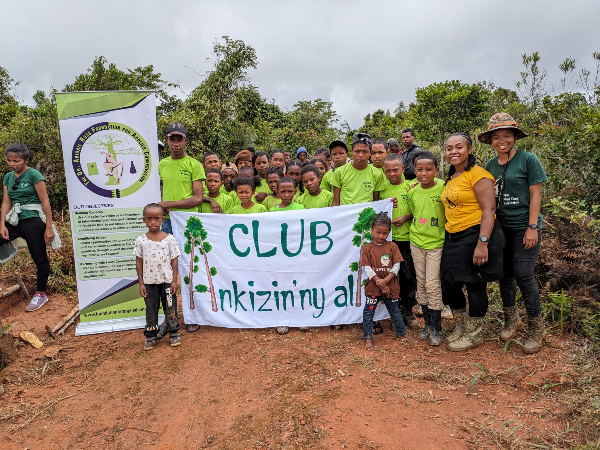 A group of people in bright green shirts stands outside with a banner that reads "Club Ankizin'ny Ala"
