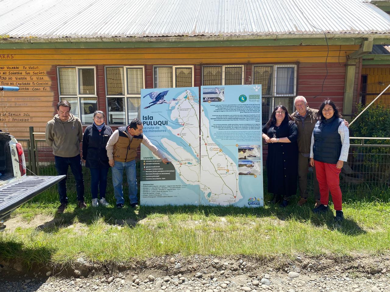 A group of people poses with a large sign displaying a picture and information about Puluqui Island