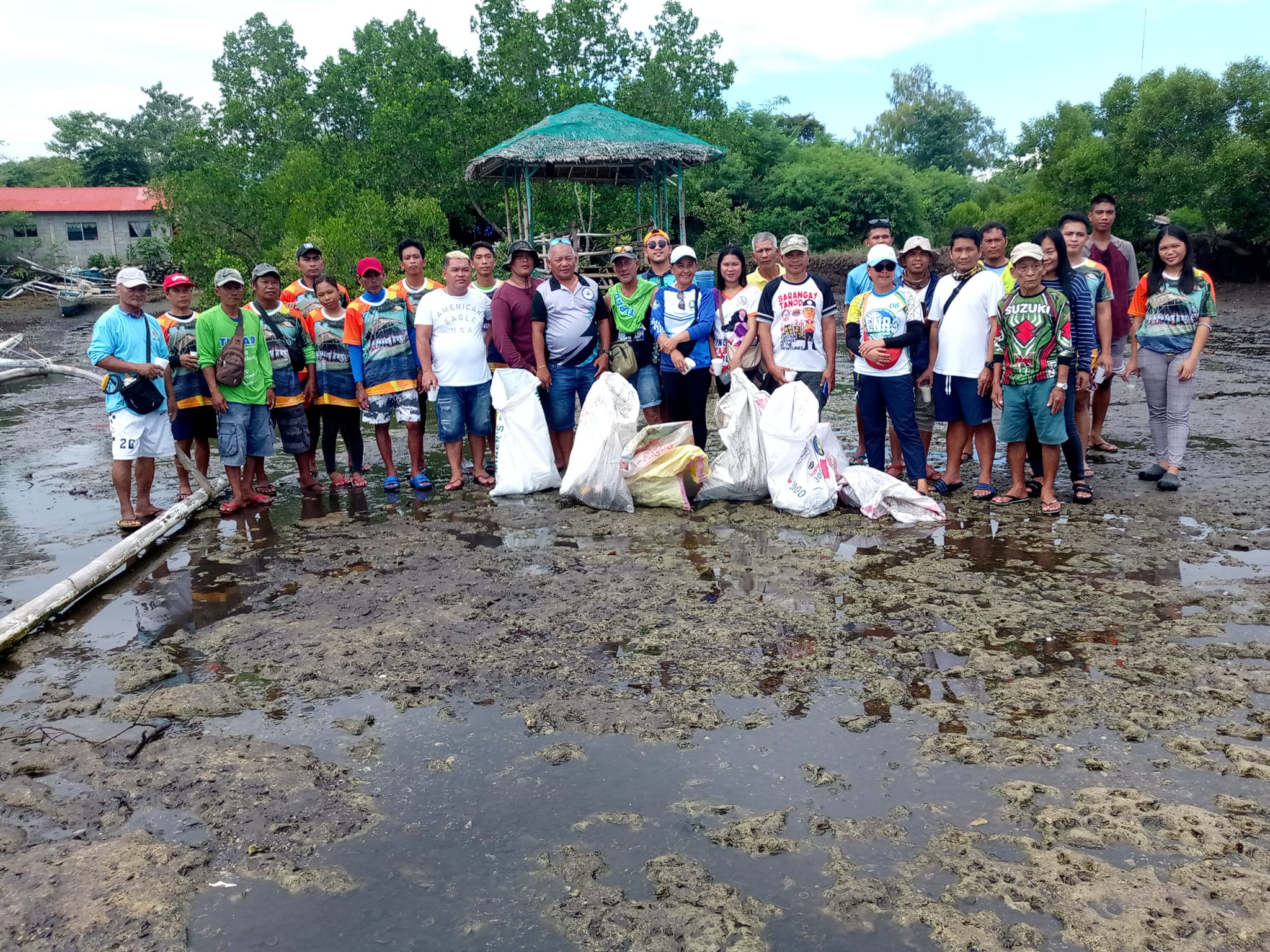 A group of people holding full trash bags stands on muddy ground in front of green trees