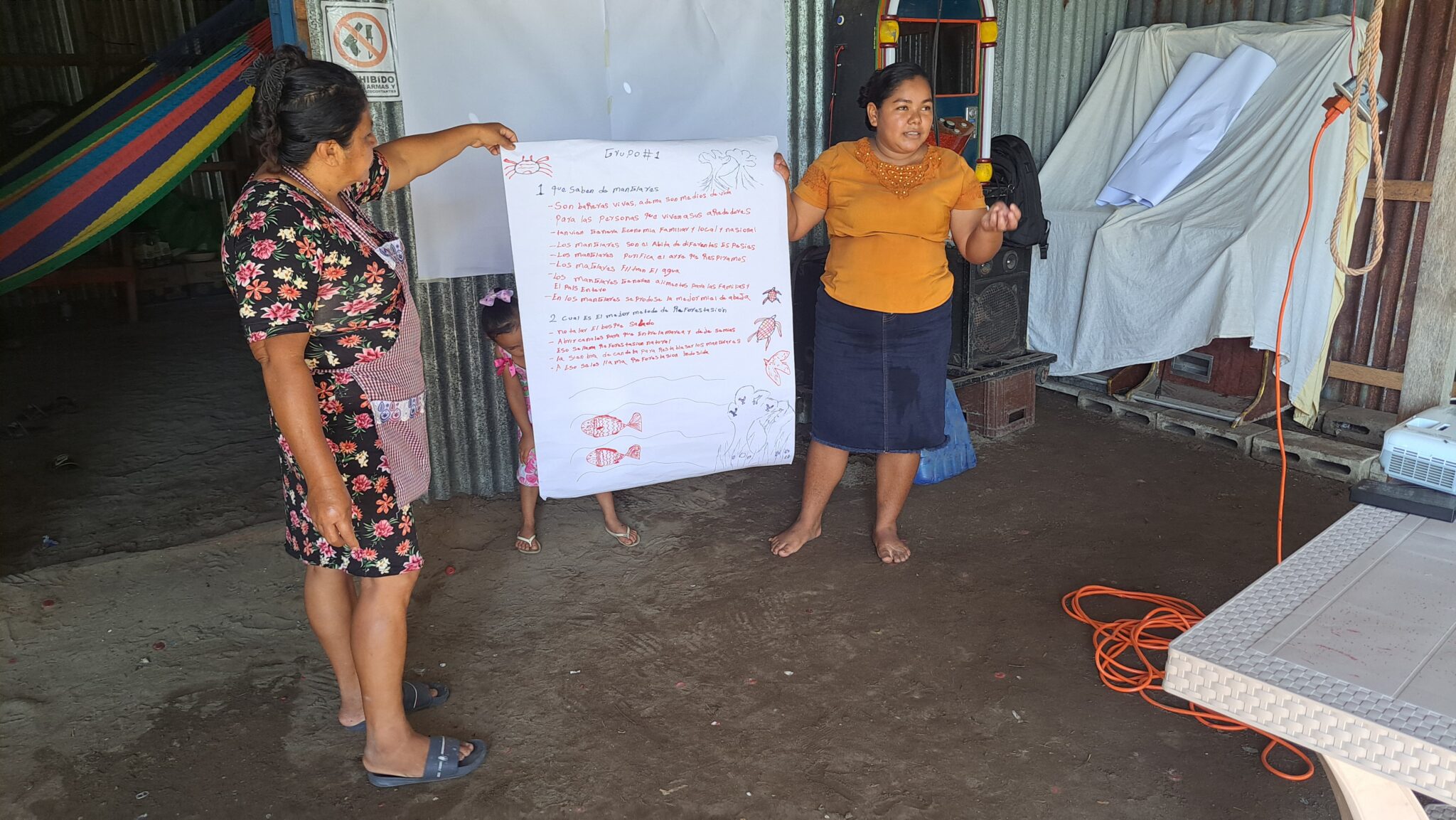 Two women holding a large informational poster with information about mangroves address a meeting