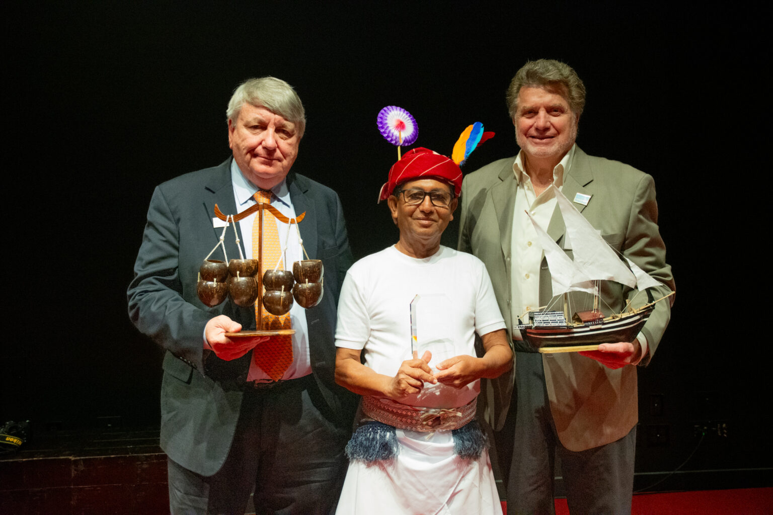 Three men stand holding model boat, trophy, and device with coconut shells