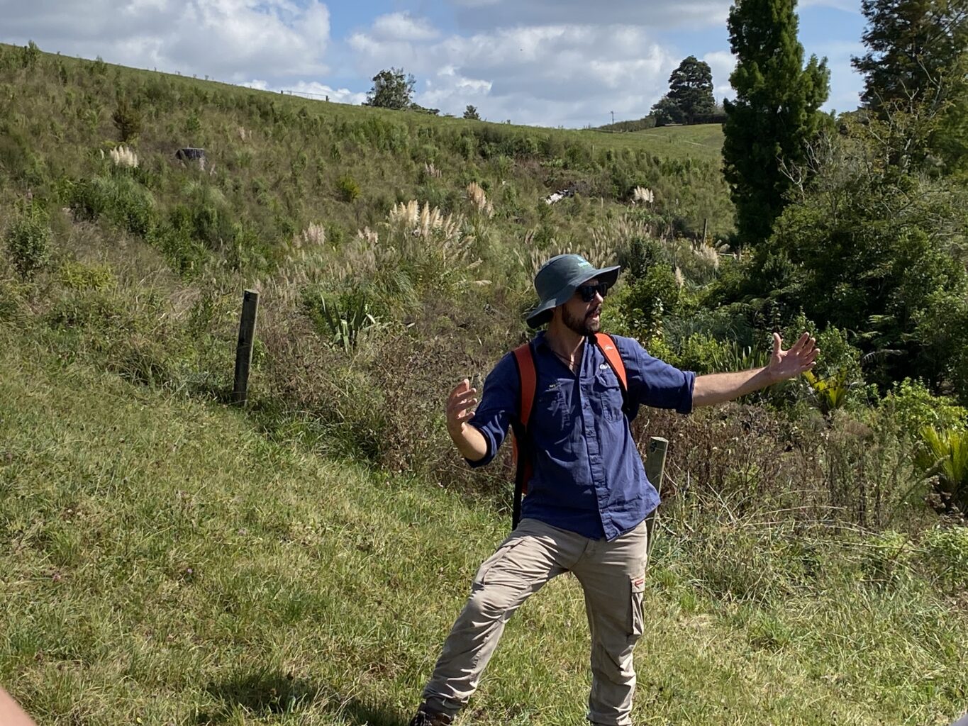 A person stands on a green grassy hillside gesturing to the landscape