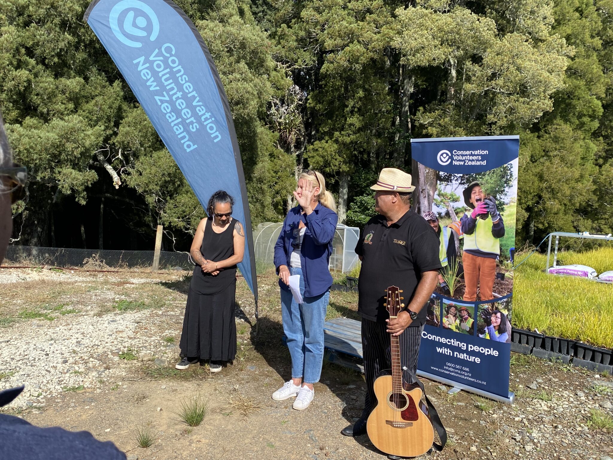 Three people, one with a guitar, stand in front of banners outside in a field