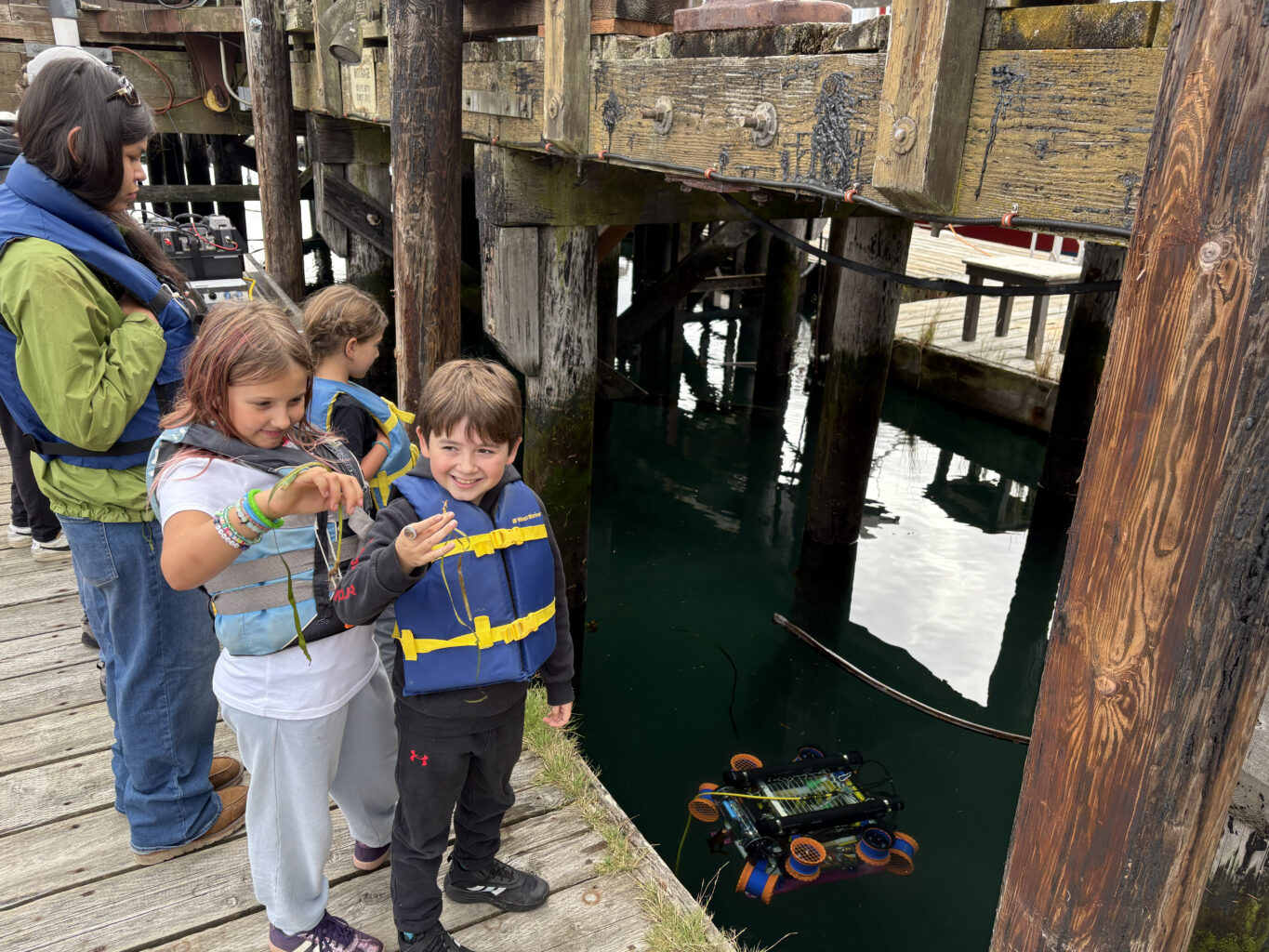 kids on pier hold seagrass shoots while robot floats in water in background