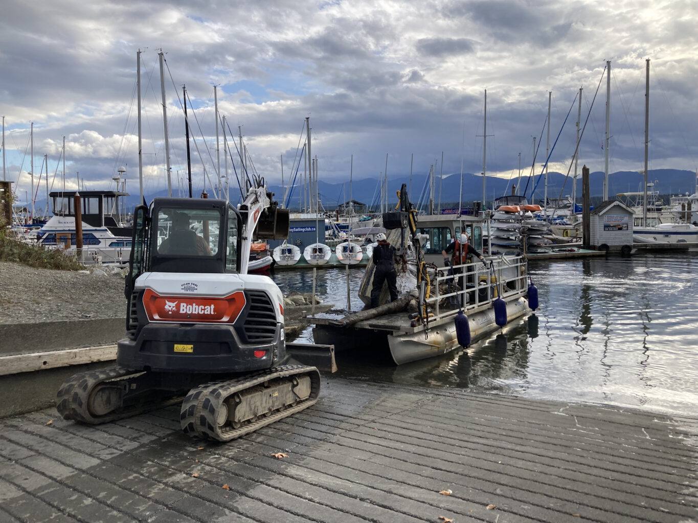 A backhoe unloads garbage from a barge on a boat ramp