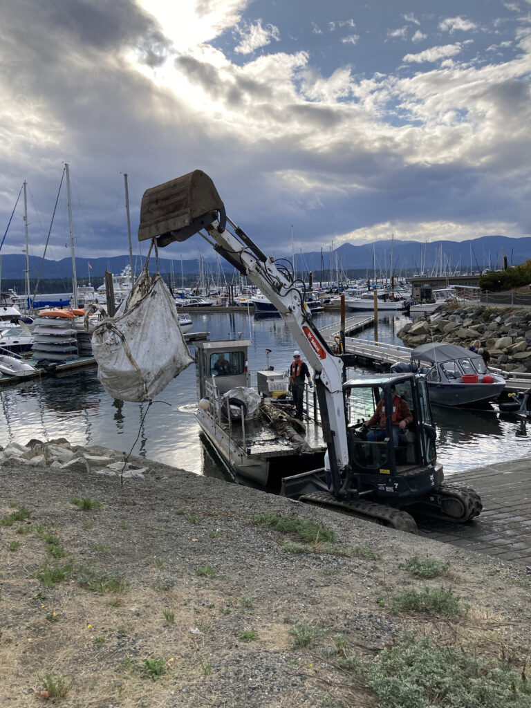A backhoe unloads garbage from a barge on a boat ramp