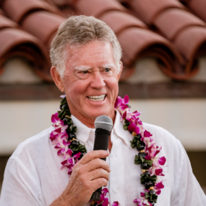 smiling man with flower garland around his neck speaks into microphone