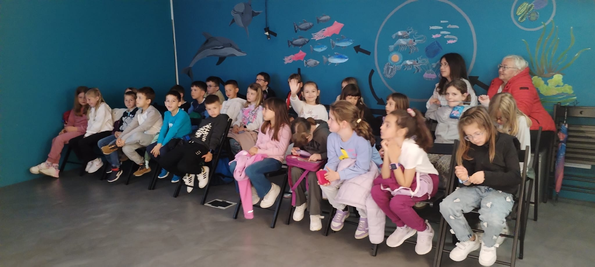 A group of kids sits in a room with an ocean mural on the wall