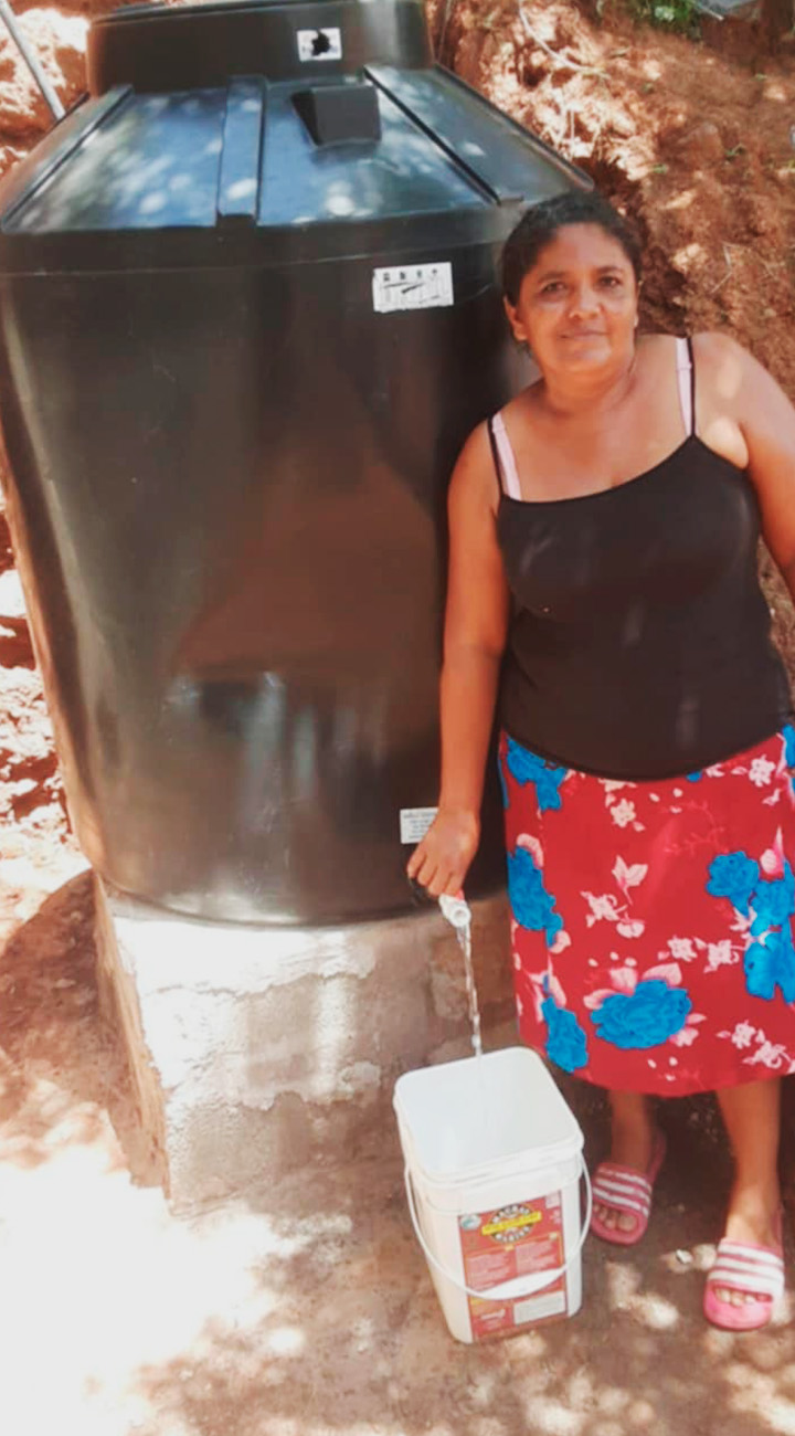 A person stands next to a large tank with water coming out of the spigot into a bucket