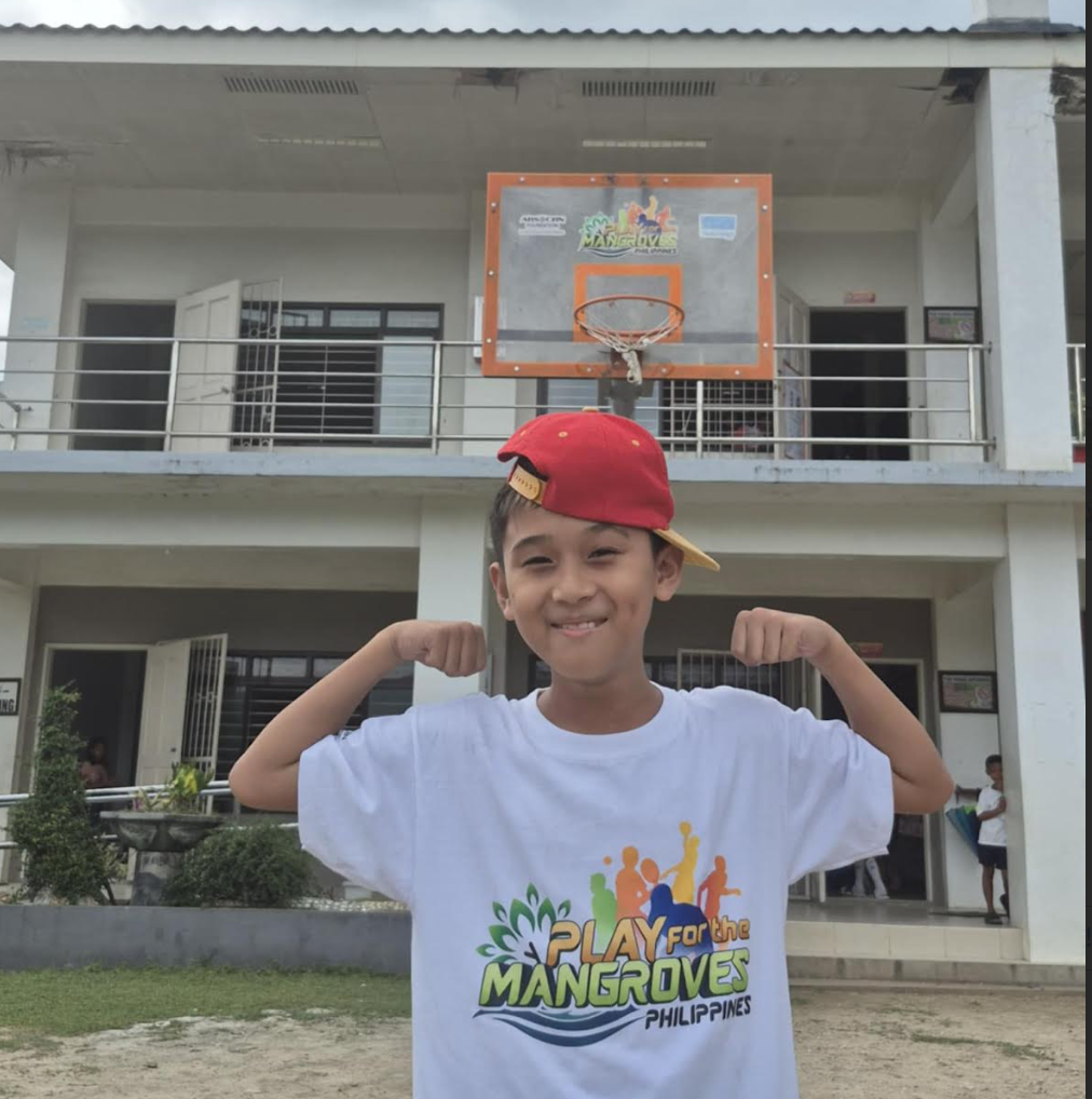 A child poses in a "strong muscle" pose in front of a basketball hoop and backboard