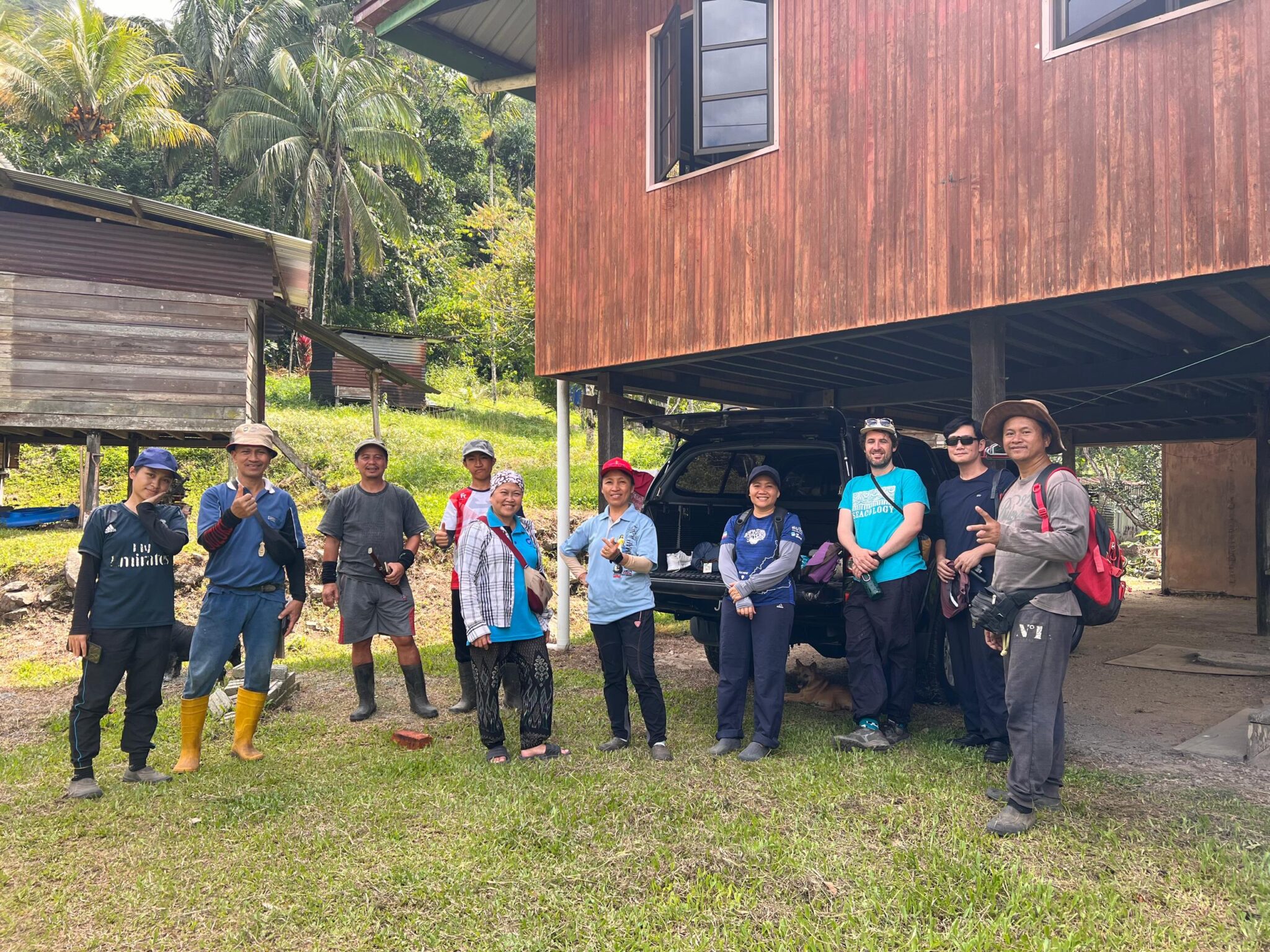 A group of people in hiking gear stand next to a raised building with palm trees in the background.