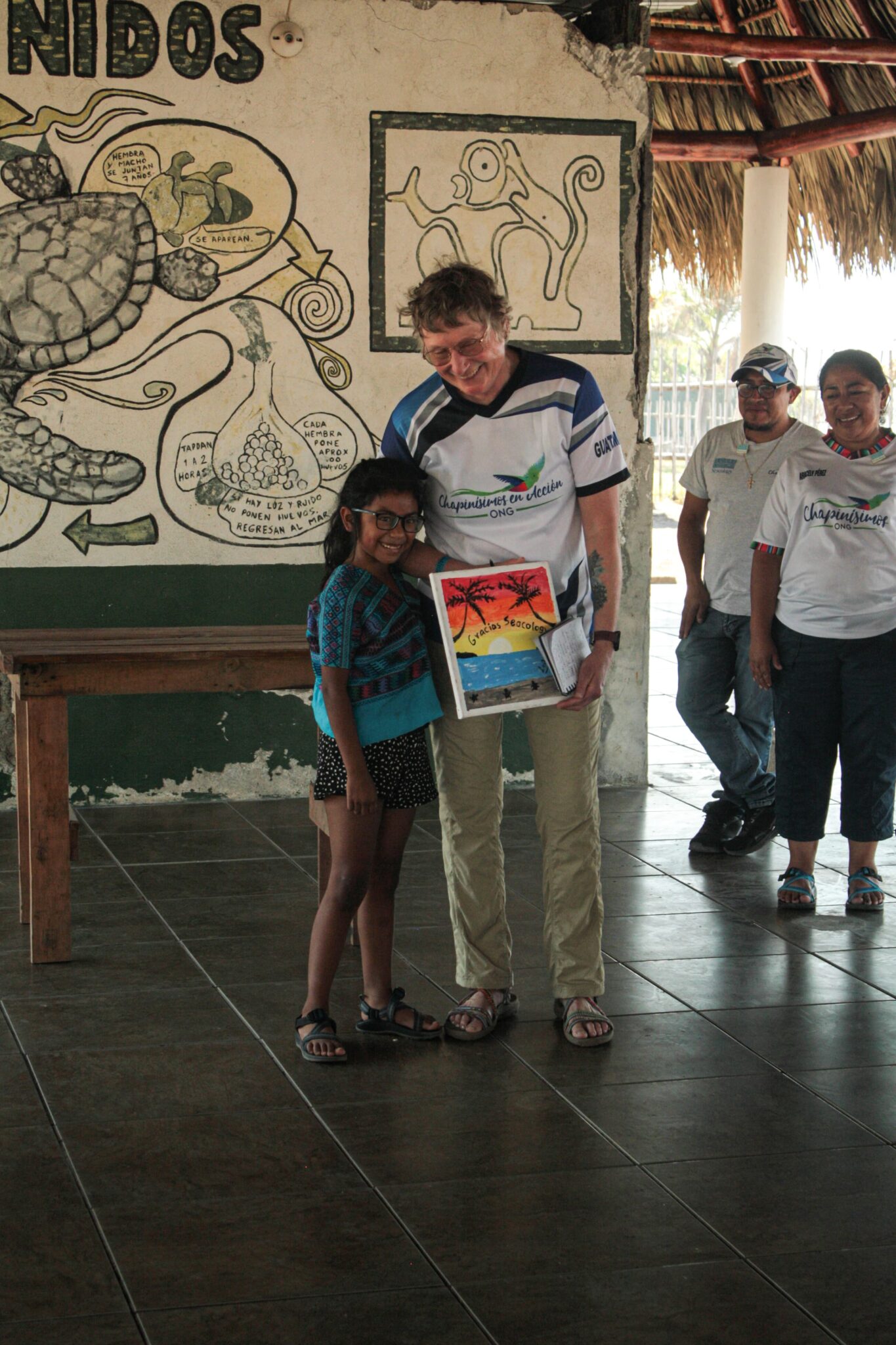 A child stands with an adult, both smiling, and holding a brightly colored painting