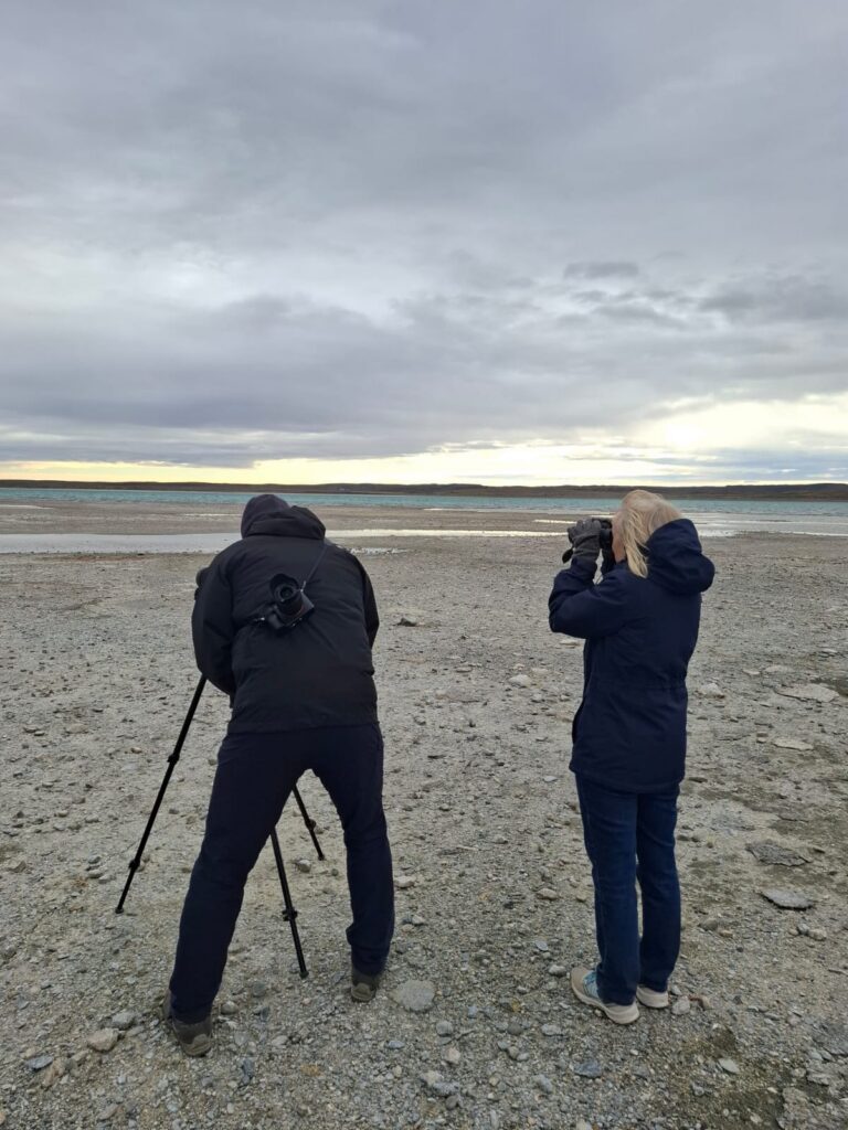 Two people look through binoculars across a mud flat towards water