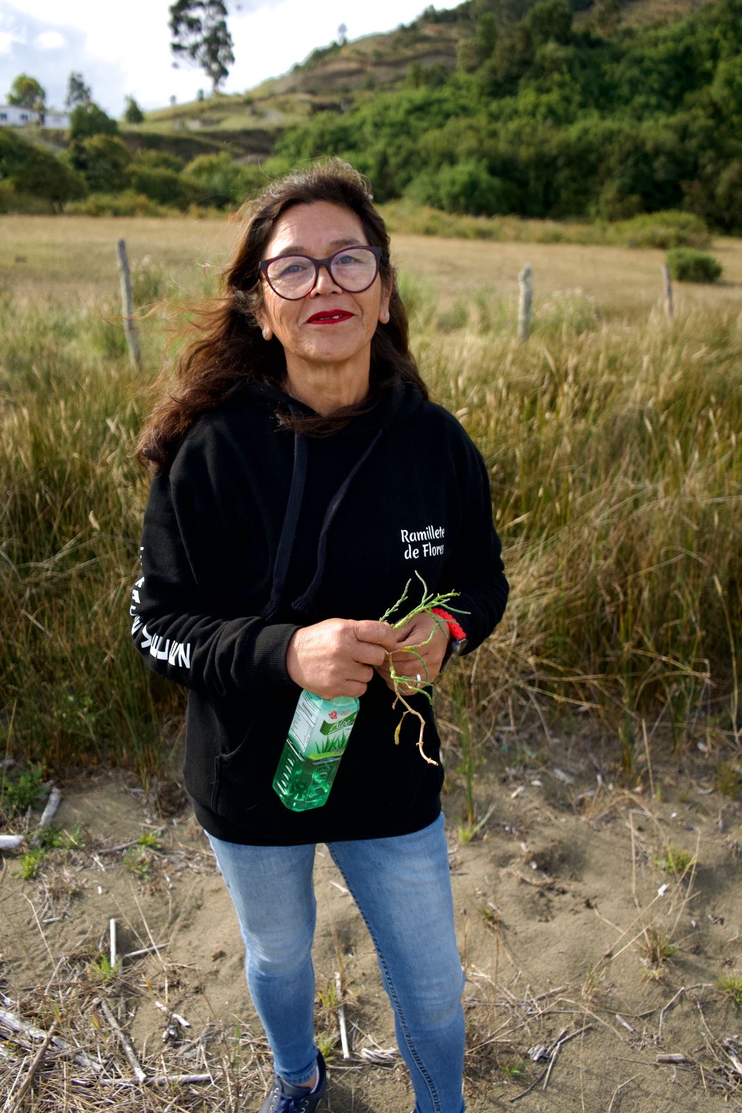 A person stands in front of a field holding a bottle and some grass